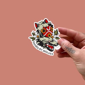 May include: A sticker depicting a wolf with a red and yellow mask, floral details, and the words "RAISED BY WOLVES." The sticker has a white border and is held by a hand against a peach background. The wolf has a detailed design with sharp teeth.
