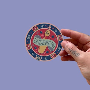 Peut inclure: Un autocollant rond avec un design rétro. Il présente une quille de bowling avec une bannière indiquant "DUDENESS". Le design comprend des cassettes, des étoiles et le mot "his". L'autocollant est tenu par une main.
