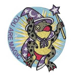 Pode incluir: Patch bordado com um sapo extravagante vestido de mago, usando um chapéu e capa roxos com detalhes de estrelas. O sapo segura uma varinha em forma de estrela. O patch é circular com as palavras "YOU ARE MAGIC" em um fundo azul claro.