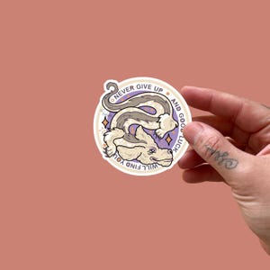 May include: A circular sticker with a cartoon-style illustration of a dog-like creature with a long, winding body in grey and cream. The sticker has the text "NEVER GIVE UP AND GOOD LUCK WILL FIND YOU" in a circular pattern around the creature.
