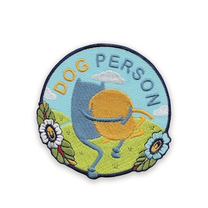 Peut inclure: Écusson brodé avec une bordure bleue. Il présente une illustration de dessin animé de deux personnages s'embrassant. Les personnages sont bleus et jaunes, sur fond de ciel bleu clair avec les mots "DOG PERSON" en jaune. L'herbe verte et les fleurs entourent les personnages.