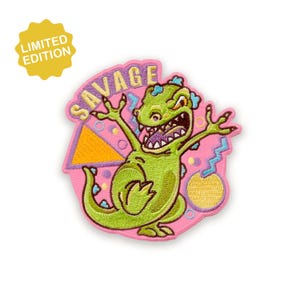 May include: A green cartoon dinosaur patch with the word "SAVAGE" in yellow lettering. The patch has a pink background with a yellow triangle, blue zigzags, and yellow circles. A yellow "LIMITED EDITION" sticker is in the upper left corner.