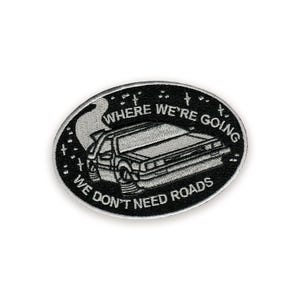 Può includere: Toppa ovale in bianco e nero con un'auto DeLorean e la scritta "WHERE WE'RE GOING WE DON'T NEED ROADS". L'auto è raffigurata con scie di fumo e circondata da stelle.