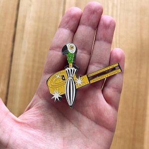 Op de afbeelding: Een kleurrijke emaille pin met een papegaaienkop, een zwart-wit gestreepte jurk en een gele meetlint. De tekst op de tape luidt "PRACTICALLY PERFECT". De pin is een uniek accessoire.