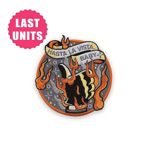 Może przedstawiać: Haftowana naszywka z okrągłym wzorem przedstawiającym czarno-żółty obraz otoczony pomarańczowymi płomieniami. Naszywka zawiera napis "HASTA LA VISTA BABY" na banerze. Różowe koło z napisem "LAST UNITS" znajduje się w lewym górnym rogu.