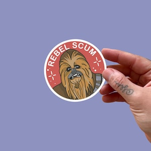 May include: A circular sticker with a cartoon depiction of Chewbacca and the words "REBEL SCUM" in a red circle. The sticker has a white border and is set against a purple backdrop.