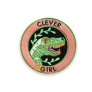 May include: Embroidered patch featuring a green dinosaur head with orange accents and a purple tongue, set against a black background. The patch is encircled by a pink ring with the words "CLEVER GIRL" in a curved font.