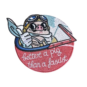 Könnte beinhalten: Ein runder gestickter Aufnäher mit einem Cartoon-Schwein, das eine Fliegerbrille und einen Schal trägt und im Cockpit den Daumen hochhält. Der rote untere Bereich enthält den weißen gestickten Text: better a pig than a fascist.
