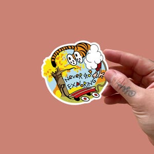 May include: A colorful sticker featuring a cartoon tiger with the text "Never Stop Exploring." The tiger is next to a red wagon, a tree, and a cloud. The sticker is round with a white border.