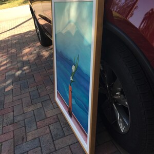 May include: A framed painting of a mountain landscape with a single white flower in a brown vase. The painting is in a gold frame and is leaning against a red car.