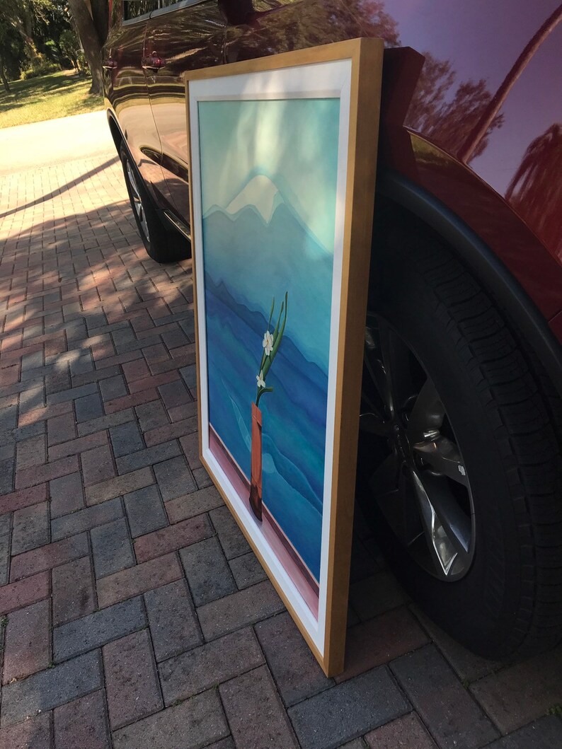 May include: A framed painting of a mountain landscape with a single white flower in a brown vase. The painting is in a gold frame and is leaning against a red car.