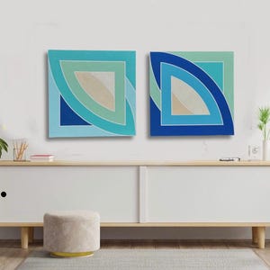 May include: Two abstract geometric paintings with blue, green, and white shapes. The paintings are hanging on a white wall above a wooden cabinet.