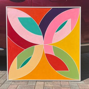 May include: Abstract geometric painting with a white frame. The painting features a colorful design with overlapping shapes in shades of pink, red, orange, yellow, green, and purple.