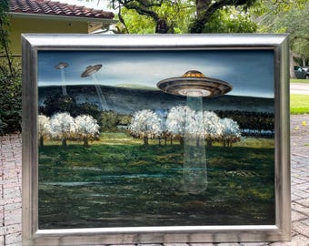 May include: A framed painting depicts a landscape with three UFOs in the sky. The foreground shows white-blossomed trees and green grass. The UFOs are silver and gold, with beams of light shining down. The background features a dark hill and a blue sky.