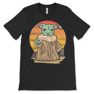 Puede incluir: Una camiseta negra con una imagen de dibujos animados de un perro vestido como un personaje de la franquicia Star Wars. El perro es verde y marr&oacute;n con una cabeza grande y orejas grandes. El perro lleva una t&uacute;nica marr&oacute;n con capucha y est&aacute; de pie frente a una puesta de sol.