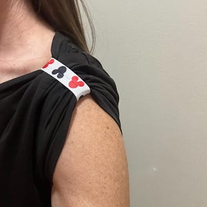 May include: Black short-sleeved top with a white band featuring red and black club suit symbols. The band is wrapped around the sleeve, adding a decorative detail to the garment. The top is made of a soft, flowing fabric.