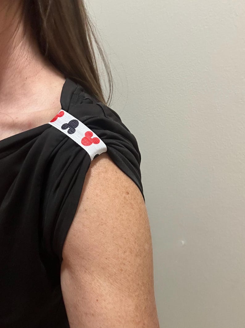 May include: Black short-sleeved top with a white band featuring red and black club suit symbols. The band is wrapped around the sleeve, adding a decorative detail to the garment. The top is made of a soft, flowing fabric.