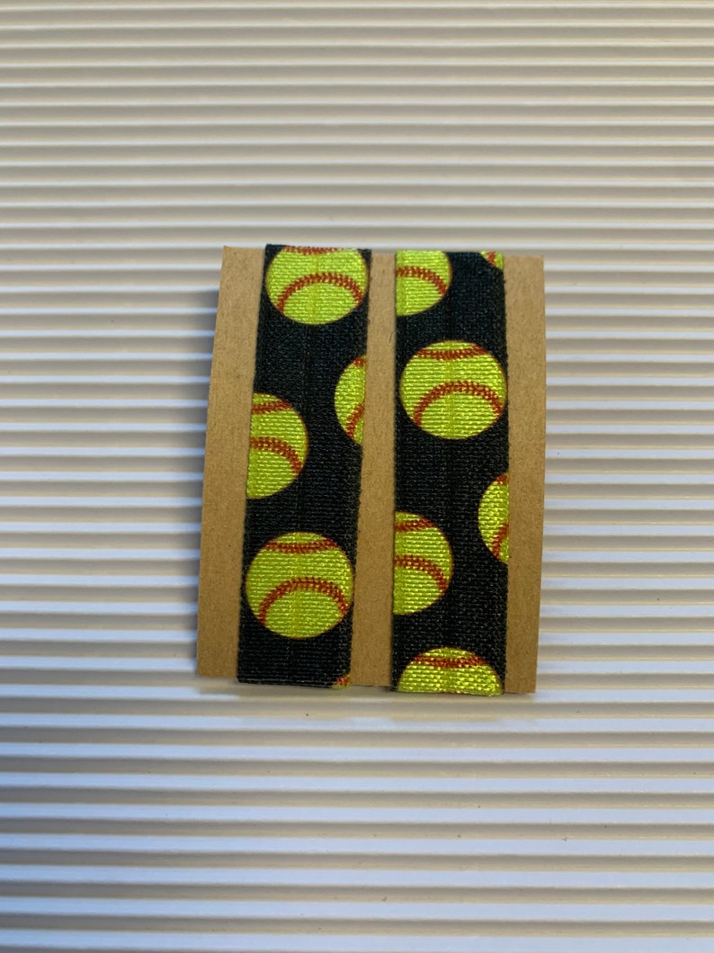 K&ouml;nnte beinhalten: Zwei schwarze Gummib&auml;nder f&uuml;r Haare mit einem sich wiederholenden Muster aus gelben Softball-Designs.