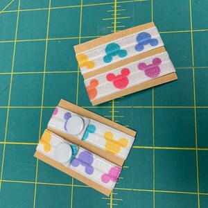 May include: Elastic bands with colorful Mickey Mouse head designs. The bands are white with the iconic silhouette in shades of pink, yellow, blue, and purple. Two sets are shown, one with snaps attached. The bands are on small cardboard rectangles.