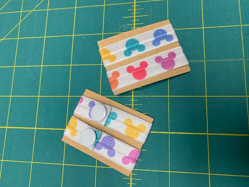 May include: Elastic bands with colorful Mickey Mouse head designs. The bands are white with the iconic silhouette in shades of pink, yellow, blue, and purple. Two sets are shown, one with snaps attached. The bands are on small cardboard rectangles.