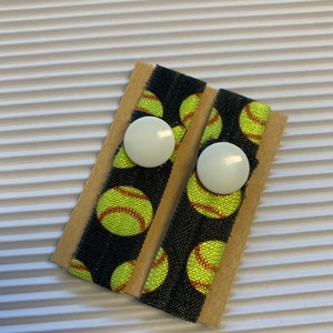 K&ouml;nnte beinhalten: Zwei schwarze Gummib&auml;nder f&uuml;r Haare mit einem wei&szlig;en Knopf und einem Muster aus gelben und roten Softball-Designs auf braunem Hintergrund.