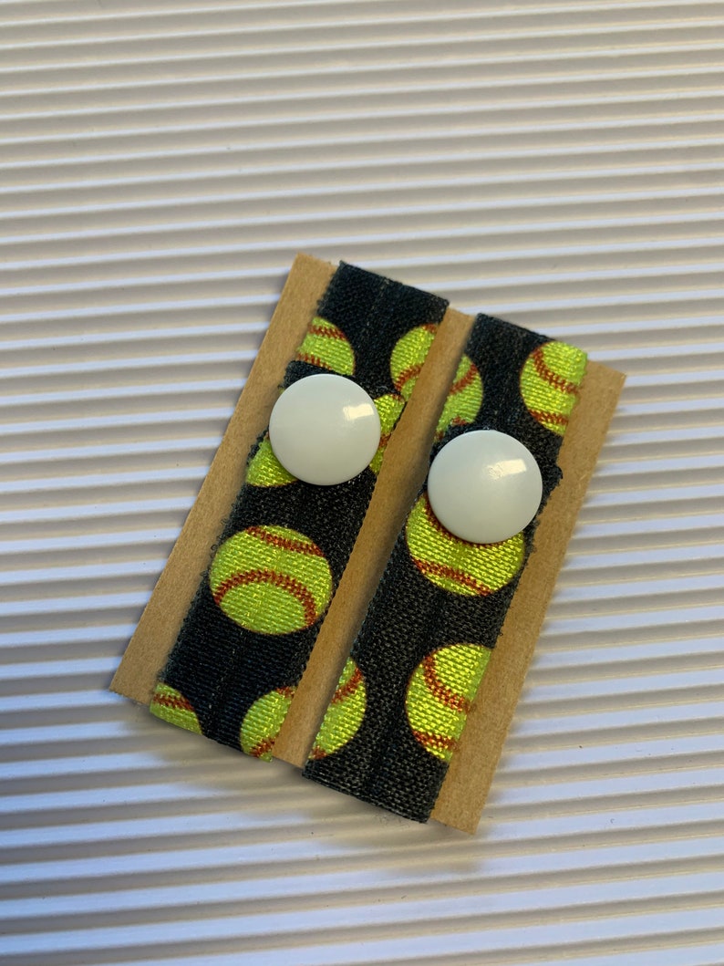 K&ouml;nnte beinhalten: Zwei schwarze Gummib&auml;nder f&uuml;r Haare mit einem wei&szlig;en Knopf und einem Muster aus gelben und roten Softball-Designs auf braunem Hintergrund.