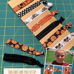 May include: A set of basketball-themed hair ties in various patterns, including basketballs, polka dots, and text. The ties are attached to a brown card with a black and white string. The image also includes a small photo collage.