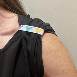 May include: Black sleeveless top with a white strap featuring a pattern of blue and yellow Mickey Mouse silhouettes. The strap is positioned over the shoulder of the top.