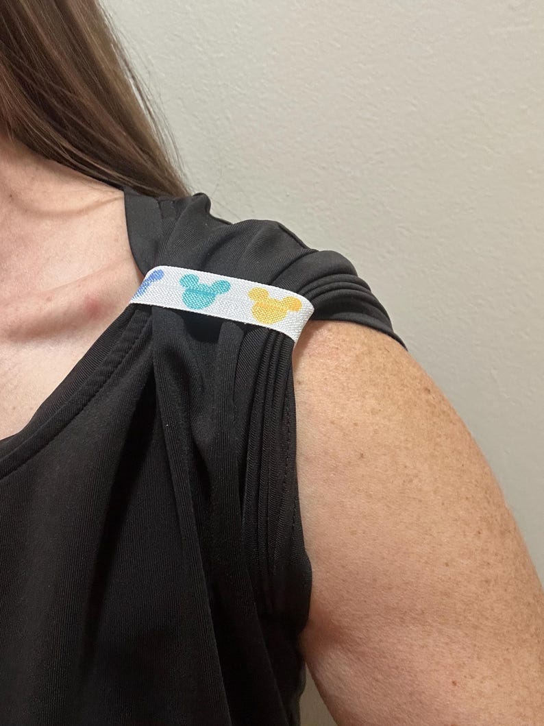 May include: Black sleeveless top with a white strap featuring a pattern of blue and yellow Mickey Mouse silhouettes. The strap is positioned over the shoulder of the top.