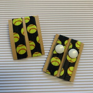 K&ouml;nnte beinhalten: Zwei Paar schwarze Gummib&auml;nder f&uuml;r Haare mit einem gelben Softball-Muster. Jedes Paar ist auf einer braunen Karte mit zwei wei&szlig;en Kn&ouml;pfen verpackt.