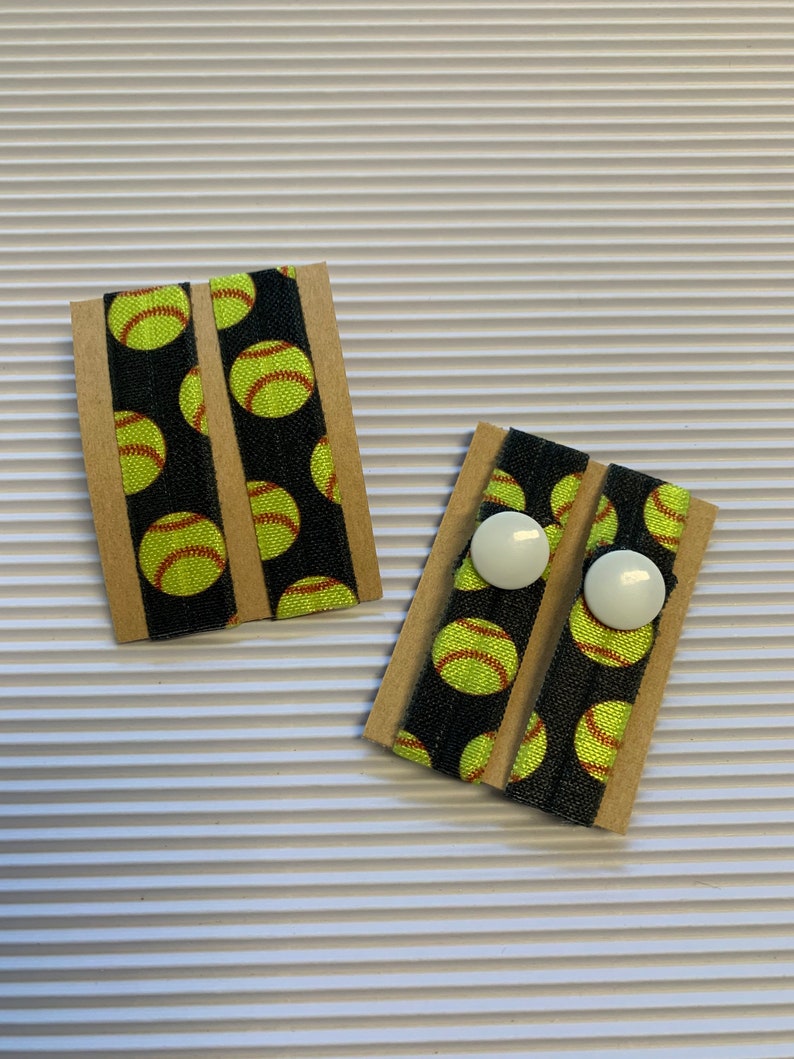K&ouml;nnte beinhalten: Zwei Paar schwarze Gummib&auml;nder f&uuml;r Haare mit einem gelben Softball-Muster. Jedes Paar ist auf einer braunen Karte mit zwei wei&szlig;en Kn&ouml;pfen verpackt.