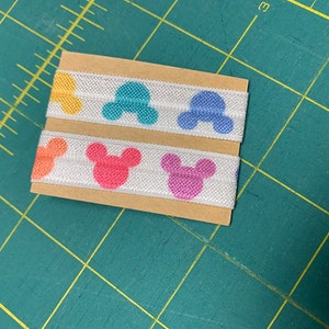 May include: Two stacks of white elastic ribbon with colorful Mickey Mouse head patterns in yellow, orange, pink, blue, and purple. The ribbon is on a brown cardboard backing, set against a green cutting mat.