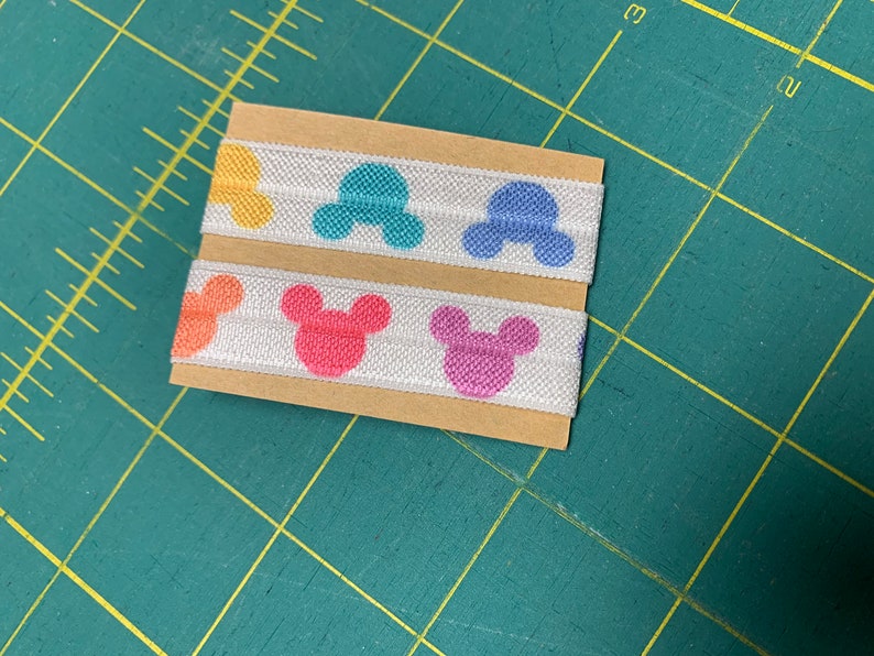 May include: Two stacks of white elastic ribbon with colorful Mickey Mouse head patterns in yellow, orange, pink, blue, and purple. The ribbon is on a brown cardboard backing, set against a green cutting mat.