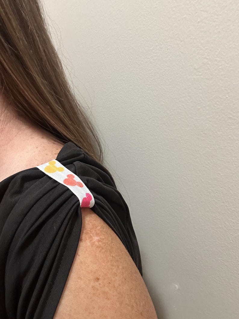 May include: Black top with a white strap featuring a repeating pattern of yellow, orange, and pink Mickey Mouse heads. The strap is sewn onto the shoulder of the top.