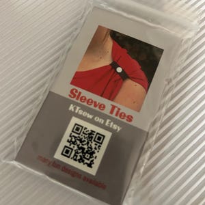 May include: A clear plastic bag containing a product card for "Sleeve Ties." The card features a photo of a black sleeve tie on a red top. Text includes "KTsew on Etsy" and a QR code, with the phrase "many fun designs available."
