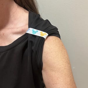 May include: Black sleeveless top with a white band featuring blue and yellow cartoon character designs. The band is sewn onto the shoulder of the top. The top has a square neckline and gathered fabric details.