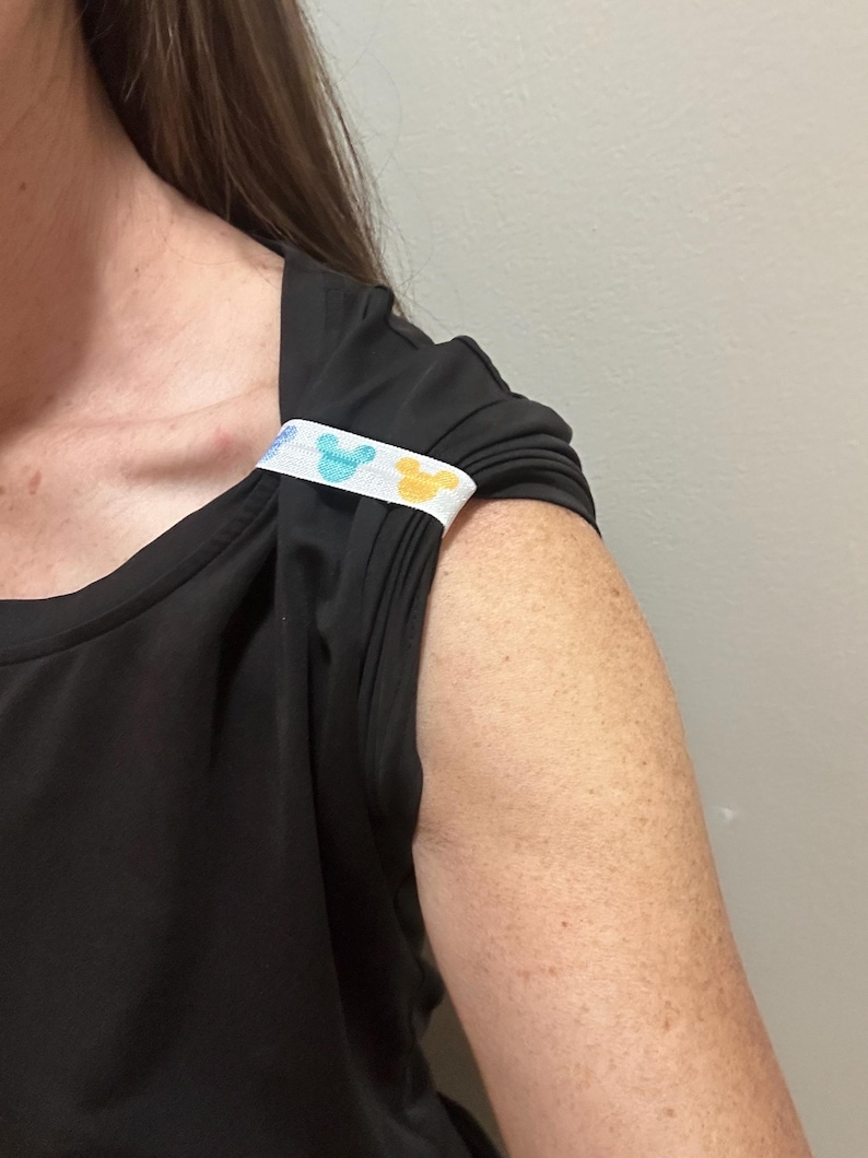 May include: Black sleeveless top with a white band featuring blue and yellow cartoon character designs. The band is sewn onto the shoulder of the top. The top has a square neckline and gathered fabric details.