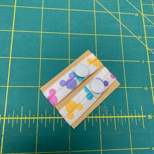 May include: Two white elastic bands with colorful Mickey Mouse head patterns, each attached to a small cardboard rectangle. Each band has a white button. The background is a green cutting mat.