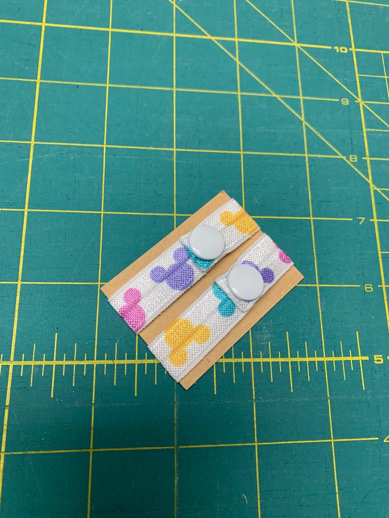 May include: Two white elastic bands with colorful Mickey Mouse head patterns, each attached to a small cardboard rectangle. Each band has a white button. The background is a green cutting mat.