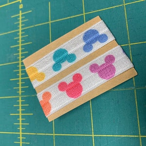 May include: Two strips of white elastic ribbon with colorful Mickey Mouse head designs. The heads are in shades of orange, yellow, teal, blue, and pink. The ribbon is mounted on a small piece of brown cardboard.