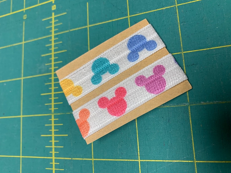 May include: Two strips of white elastic ribbon with colorful Mickey Mouse head designs. The heads are in shades of orange, yellow, teal, blue, and pink. The ribbon is mounted on a small piece of brown cardboard.