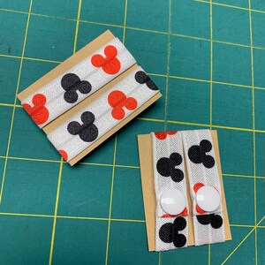 May include: Two sets of elastic hair ties featuring a repeating pattern of black and red Mickey Mouse silhouettes on a white background. Each set is attached to a small cardboard backing. One set has white buttons.