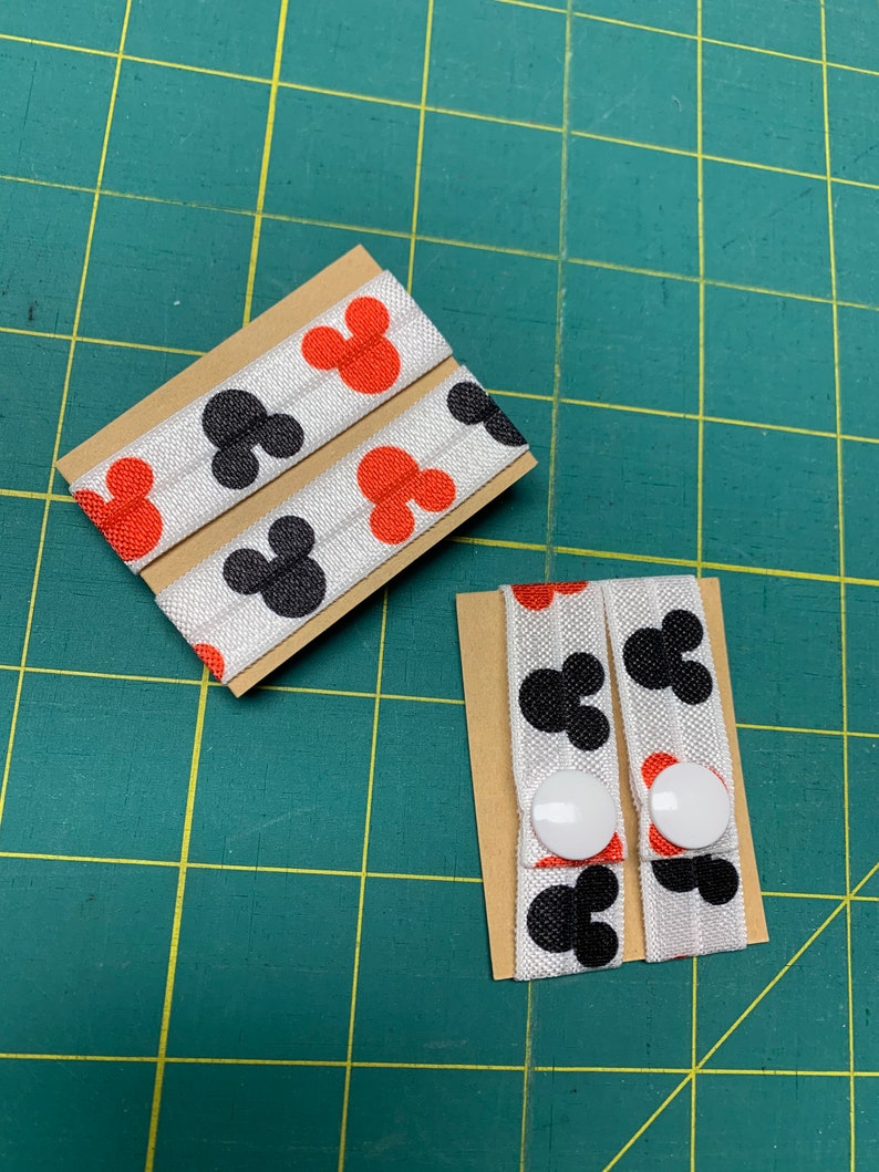 May include: Two sets of elastic hair ties featuring a repeating pattern of black and red Mickey Mouse silhouettes on a white background. Each set is attached to a small cardboard backing. One set has white buttons.