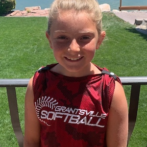 May include: A red and black softball jersey with the text "GRANTSVILLE SOFTBALL" in white. The jersey is sleeveless and has a camouflage pattern. The person is smiling and has blonde hair.