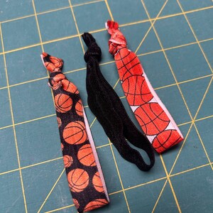 May include: Three hair ties on a green surface. Two ties feature a basketball pattern with orange and black basketballs on a black background. The third tie is solid black. The ties are knotted at the top.