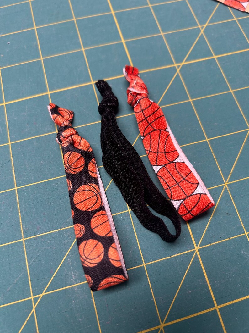 May include: Three hair ties on a green surface. Two ties feature a basketball pattern with orange and black basketballs on a black background. The third tie is solid black. The ties are knotted at the top.