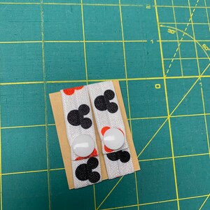 May include: Two white fabric button straps featuring black Mickey Mouse head designs and red accents. Each strap has a white button. The straps are on a small piece of cardboard, against a green cutting mat.