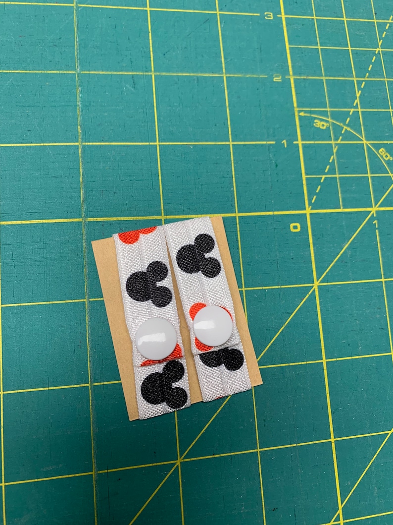 May include: Two white fabric button straps featuring black Mickey Mouse head designs and red accents. Each strap has a white button. The straps are on a small piece of cardboard, against a green cutting mat.