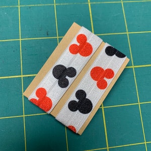 May include: Two white elastic hair ties featuring a repeating pattern of red and black Mickey Mouse-shaped figures. The hair ties are displayed on a small piece of brown card, set against a green grid background.