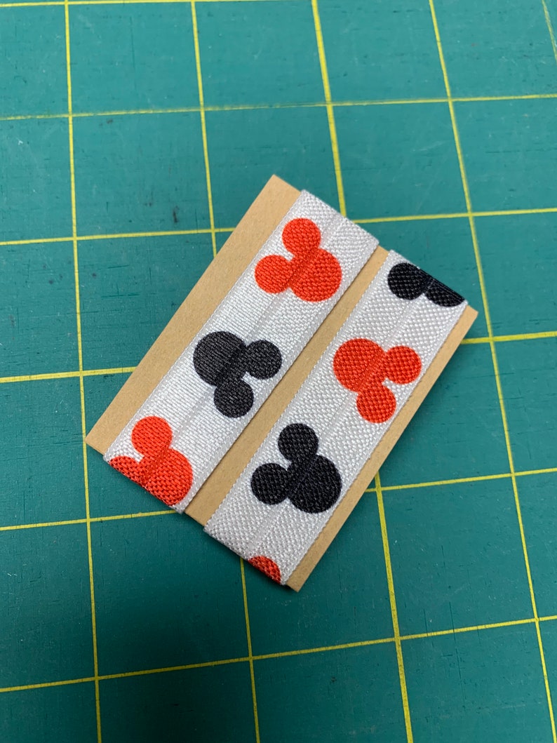May include: Two white elastic hair ties featuring a repeating pattern of red and black Mickey Mouse-shaped figures. The hair ties are displayed on a small piece of brown card, set against a green grid background.